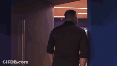 A Man In A Black Shirt Is Walking Through A Doorway GIF