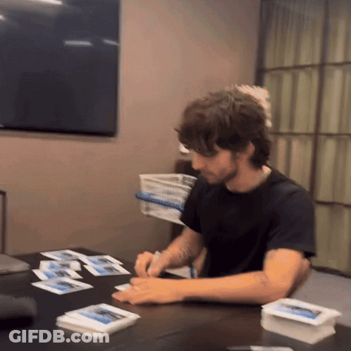 A Man In A Black Shirt Sits At A Table With A Bunch Of Cards On It GIF
