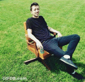 A Man In A Black Shirt Sits In A Wooden Chair In The Grass GIF