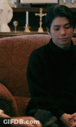 A Man In A Black Shirt Sits On A Couch In Front Of A Cross GIF