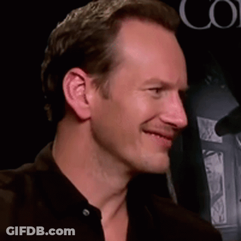 A Man In A Black Shirt Smiles In Front Of A Sign That Says Co GIF