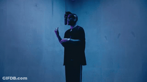 A Man In A Black Shirt Stands In Front Of A Blue Wall Holding A Microphone GIF