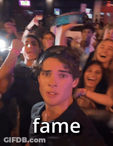 A Man In A Black Shirt Stands In Front Of A Crowd With The Word Fame Written On His Face GIF