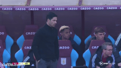 A Man In A Black Shirt Stands In Front Of A Row Of Purple And Blue Seats That Say Cazoo GIF