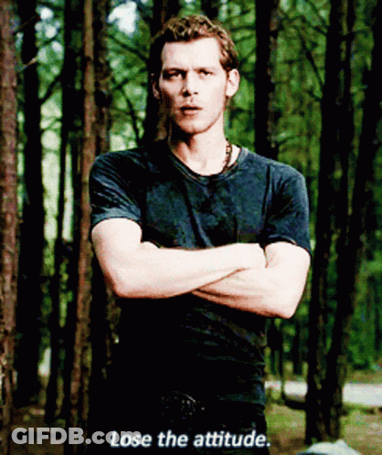 A Man In A Black Shirt Stands In The Woods With His Arms Crossed And Says " Lose The Attitude " GIF