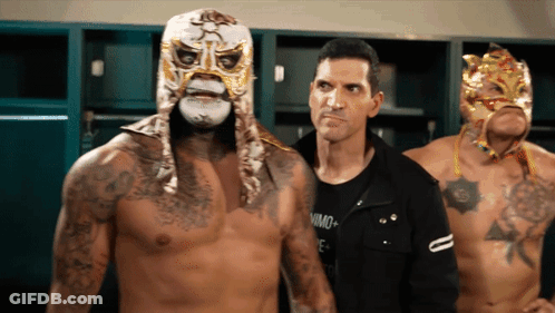 A Man In A Black Shirt Stands Next To A Man Wearing A Mask GIF