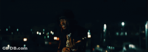 A Man In A Black Shirt With A Skull On It Is Standing In The Dark GIF
