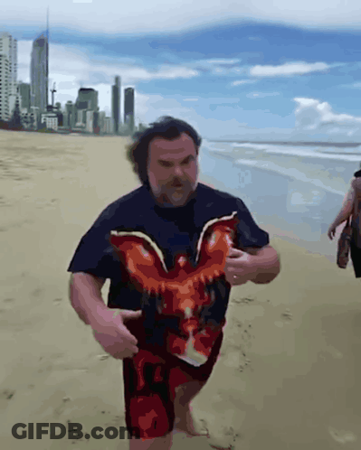 A Man In A Black Shirt With An Eagle On It Is Walking On The Beach GIF