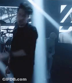 A Man In A Black Shirt With The Word Bas On The Back Is Standing In A Dark Room . GIF
