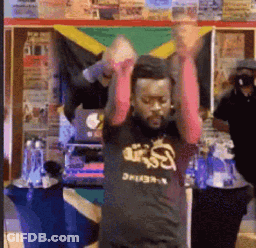 A Man In A Black Shirt With The Word Reggae On It Is Dancing In Front Of A Jamaican Flag . GIF