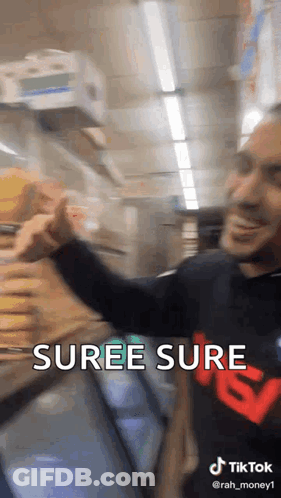 A Man In A Black Shirt With The Word Suree Sure On It Is Standing In Front Of A Refrigerator In A Store . GIF