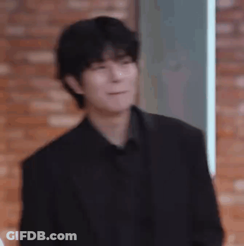 A Man In A Black Suit And Black Shirt Is Making A Funny Face While Standing In Front Of A Brick Wall . GIF
