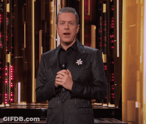 A Man In A Black Suit Holds A Microphone In Front Of A Gold Wall GIF