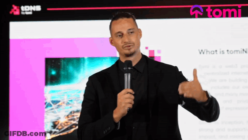 A Man In A Black Suit Holds A Microphone In Front Of A Screen That Says Tomi GIF