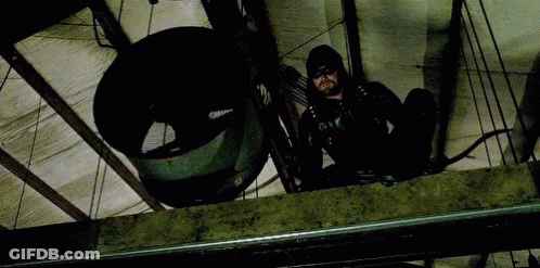 A Man In A Black Suit Is Holding A Bow And Arrow While Sitting On A Ledge . GIF