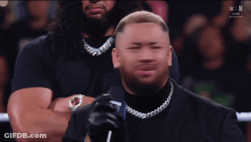 A Man In A Black Suit Is Holding A Microphone In Front Of A Crowd That Is Watching A Live Wrestling Event GIF