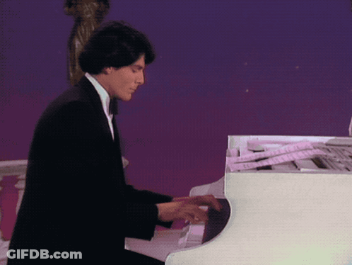 A Man In A Black Suit Is Playing A White Grand Piano GIF