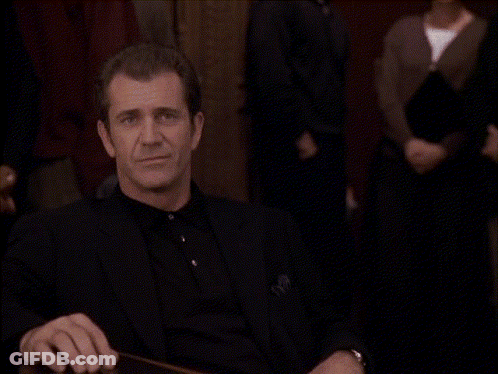 A Man In A Black Suit Is Sitting At A Table Making A Funny Face GIF