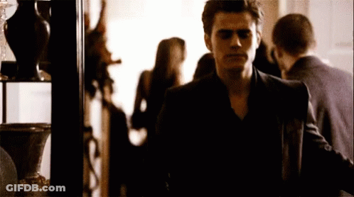 A Man In A Black Suit Is Standing In A Room With People Behind Him GIF