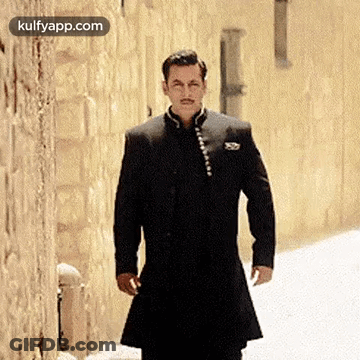 A Man In A Black Suit Is Standing In Front Of A Brick Wall . GIF