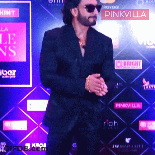 A Man In A Black Suit Is Standing In Front Of A Pinkvilla Sign GIF