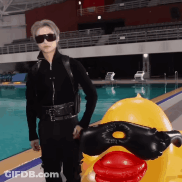 A Man In A Black Suit Is Standing Next To A Yellow Rubber Duck GIF