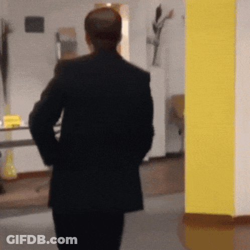 A Man In A Black Suit Is Walking In A Hallway With His Hands In His Pockets GIF