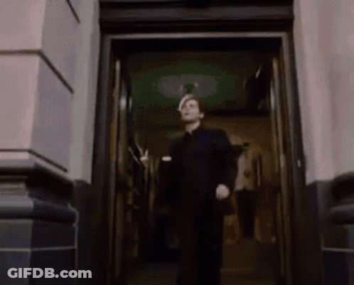 A Man In A Black Suit Is Walking Out Of A Doorway GIF