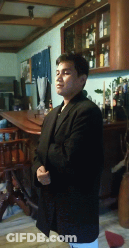 A Man In A Black Suit Stands In Front Of A Shelf Full Of Bottles GIF