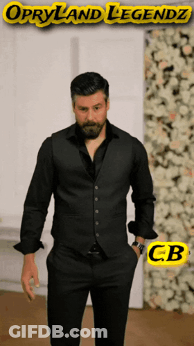 A Man In A Black Suit Stands In Front Of A Wall Of Flowers With Opryland Legendz Written On The Bottom GIF