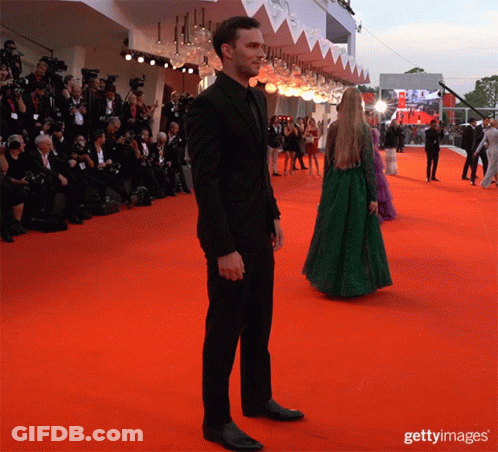 A Man In A Black Suit Stands On A Red Carpet With A Woman In A Green Dress Standing Behind Him GIF