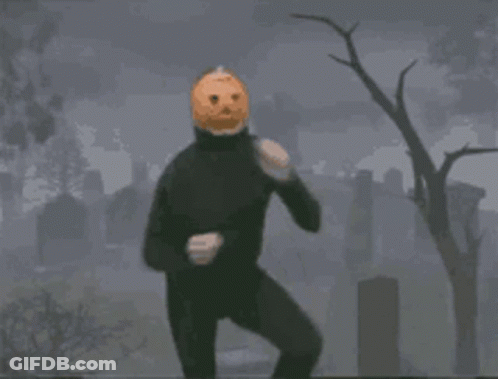 A Man In A Black Suit With A Pumpkin On His Head Is Standing In A Cemetery . GIF
