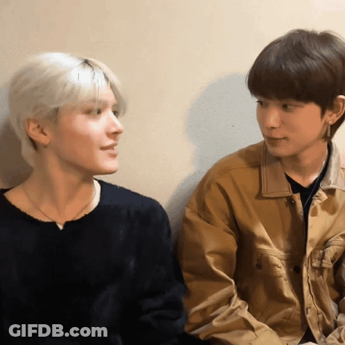 A Man In A Black Sweater And A Man In A Tan Jacket Look At Each Other GIF