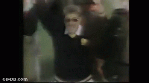 A Man In A Black Sweater And Sunglasses Is Dancing In Front Of A Crowd Of People . GIF