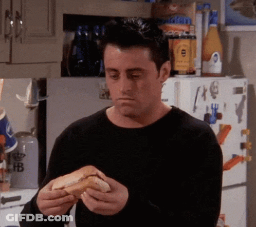 A Man In A Black Sweater Is Eating A Hot Dog In Front Of A Refrigerator In A Kitchen . GIF