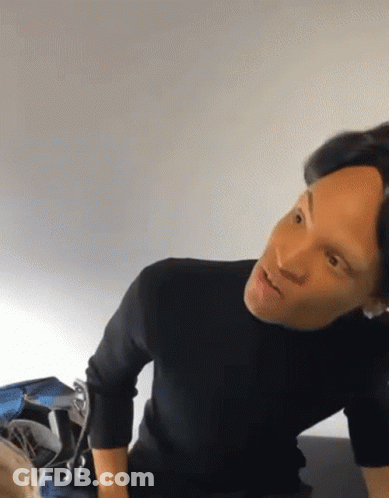 A Man In A Black Sweater Is Sitting On A Bed And Looking At Something GIF