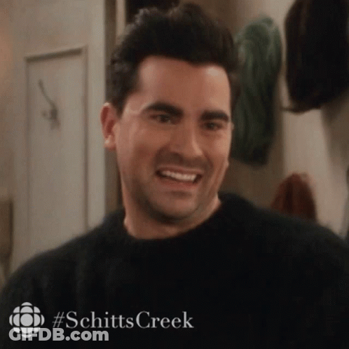 A Man In A Black Sweater Is Smiling In A Room With A Sign That Says Schitts Creek . GIF