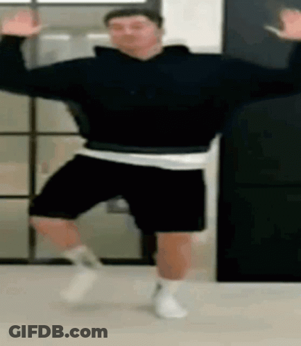 A Man In A Black Sweatshirt And Black Shorts Is Dancing In A Room . GIF