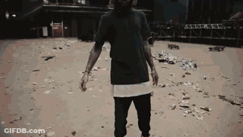 A Man In A Black T-Shirt Is Standing In A Dirt Area GIF