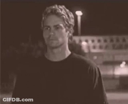 A Man In A Black T-Shirt Is Standing In Front Of A Building At Night . GIF