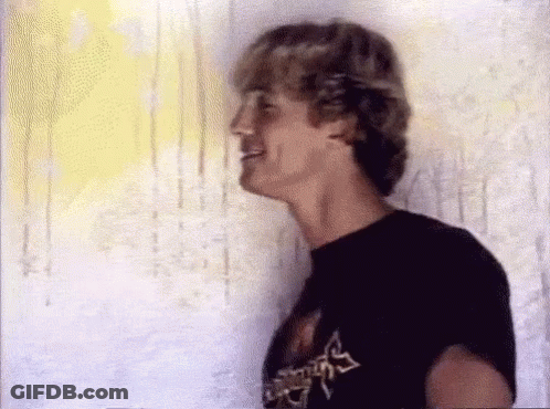 A Man In A Black T-Shirt Is Standing In Front Of A Wall And Smiling . GIF