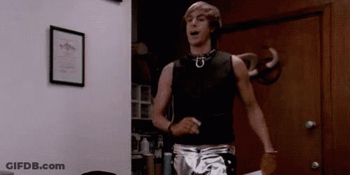 A Man In A Black Tank Top And Silver Shorts Is Standing In Front Of A Door . GIF