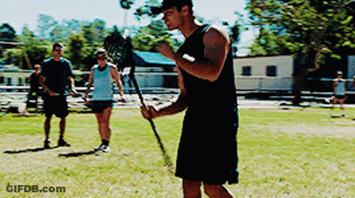 A Man In A Black Tank Top Is Holding A Lacrosse Stick In A Field GIF