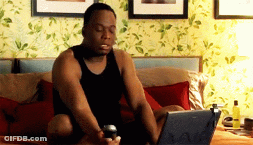 A Man In A Black Tank Top Is Sitting On A Bed Using A Laptop GIF