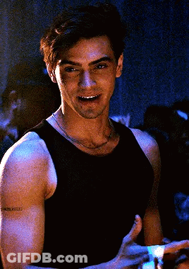 A Man In A Black Tank Top Is Smiling And Holding A Remote Control In His Hand . GIF