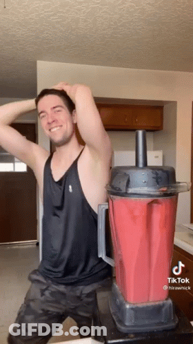 A Man In A Black Tank Top Is Standing Next To A Red Blender GIF
