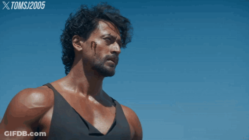 A Man In A Black Tank Top Stands In Front Of A Blue Sky With Toms 2005 Written On The Bottom Right GIF