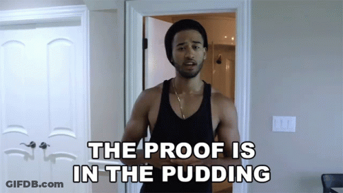 A Man In A Black Tank Top Stands In Front Of A Door With The Words The Proof Is In The Pudding Above Him GIF