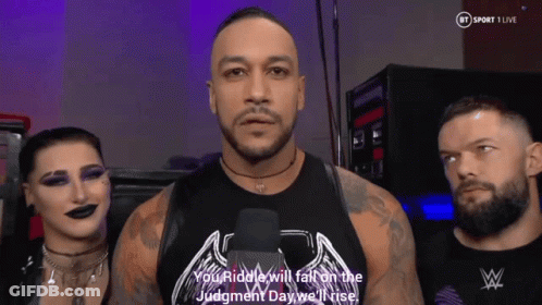 A Man In A Black Tank Top With The Word Riddle On It Stands Next To Two Other Men GIF