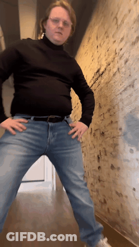 A Man In A Black Turtleneck And Blue Jeans Stands With His Hands In His Pockets GIF